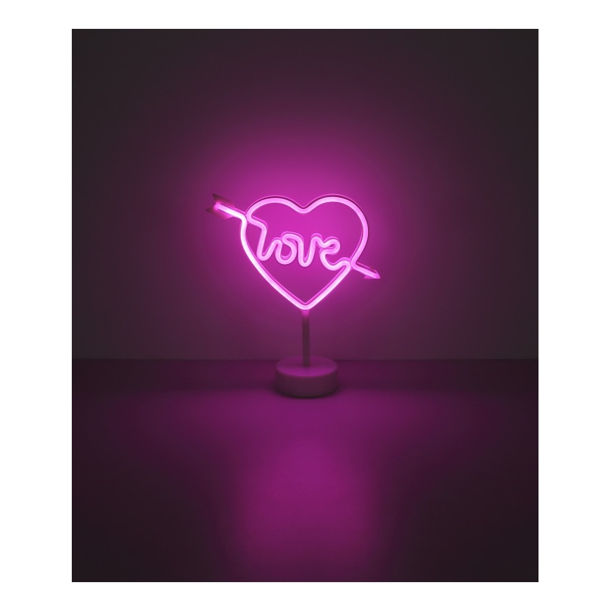 Neonové srdce s nápisom „love“ prebodnuté šípom svieti na bielom stojane. Svieti ružovým svetlom na tmavom podklade.