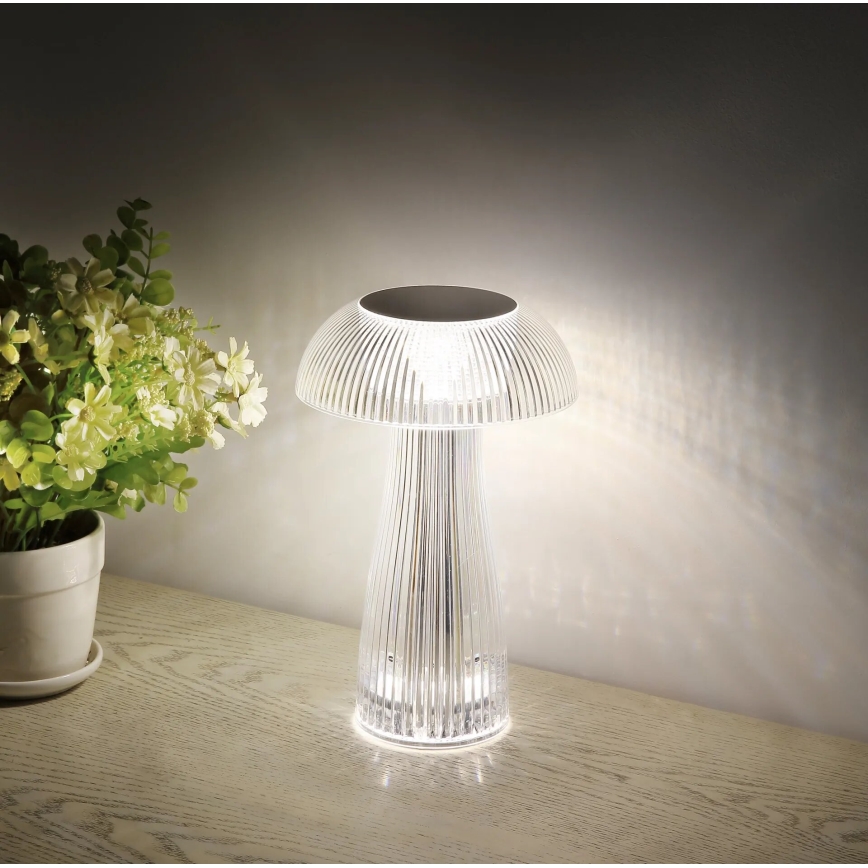 Stolová lampa v tvare huby svieti. Je umiestnená na drevenom povrchu vedľa kvetináča s kvetmi. Lampa je priehľadná s vertikálnymi drážkami.