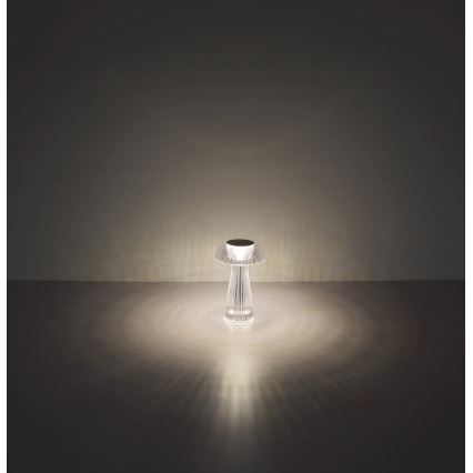 Stolná lampa svieti. Jej telo je priehľadné a má tvar stopky s rozširujúcim sa vrchným diskom. Svieti na tmavej podlahe, svetlo sa rozptyľuje do kruhu.