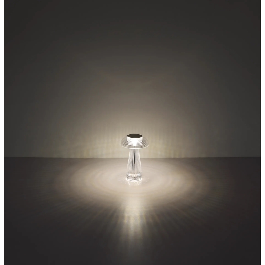 Stolná lampa svieti. Jej telo je priehľadné a má tvar stopky s rozširujúcim sa vrchným diskom. Svieti na tmavej podlahe, svetlo sa rozptyľuje do kruhu.