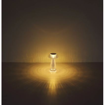 Stolová lampa s tvarom huby svieti. Svieti teplým žltým svetlom, ktoré sa odráža na podlahe. Lampa je umiestnená na tmavej podlahe pred svetlou stenou.