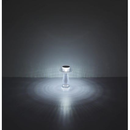 Stolová lampa svieti. Jej tvar pripomína hubu, je z priehľadného materiálu. Svietiace svetlo vytvára na podlahe svetelné kruhy.