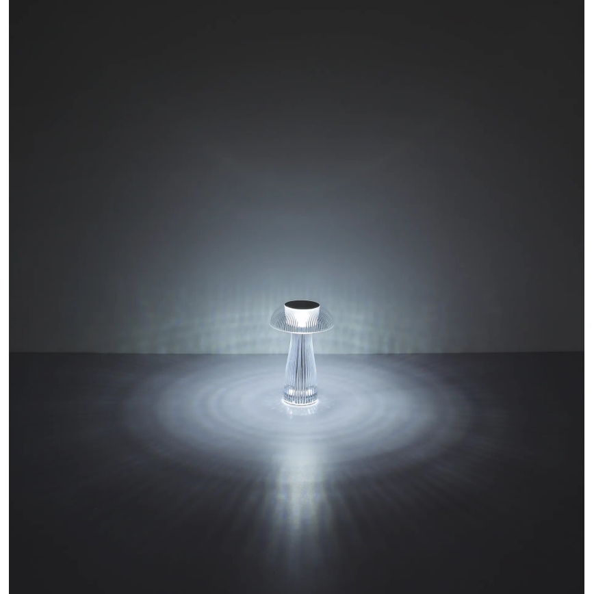 Stolová lampa svieti. Jej tvar pripomína hubu, je z priehľadného materiálu. Svietiace svetlo vytvára na podlahe svetelné kruhy.