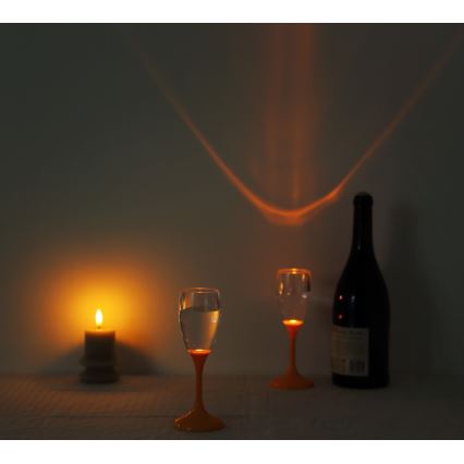 Dve poháre s vodou a oranžovými stopkami svietia zospodu. Vedľa nich je zapálená sviečka a fľaša vína. V pozadí je na stene viditeľný odraz svetla.