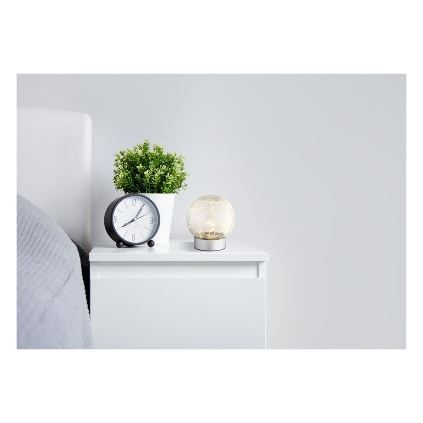 Guľová lampa s vnútorným osvetlením stojí na nočnom stolíku. Vedľa nej je budík a kvetináč s rastlinou. Lampa je umiestnená v spálni.