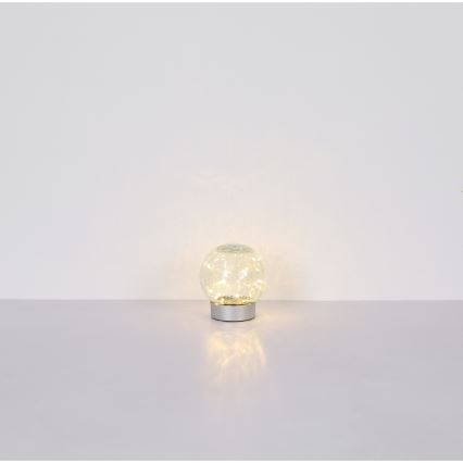 Svietiaca guľová lampa s LED diódami vnútri sklenenej gule na striebornom podstavci svieti na svetlom povrchu.