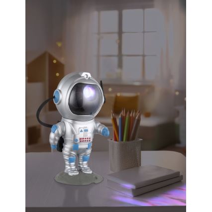 Figurína astronauta svieti a premietne svetlo na stôl. Stojí na stole vedľa pohára s ceruzkami a kníh. Svetlo projektuje farebné odlesky na knihy.