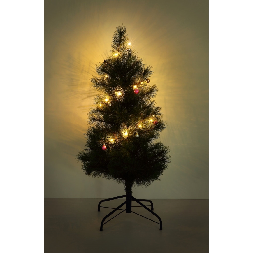 Vianočný stromček s teplými svetielkami a ozdobami stojí na čiernom stojane pred svetlou stenou. Stromček je umelý, ihličnatý.