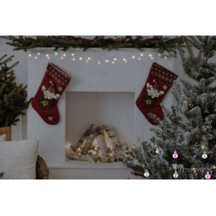 Vianočný stromček je zdobený malými guľkami a svetielkami. Za ním je krb s vianočnými pančuchami a girlandou. V miestnosti je tiež vianočná atmosféra.