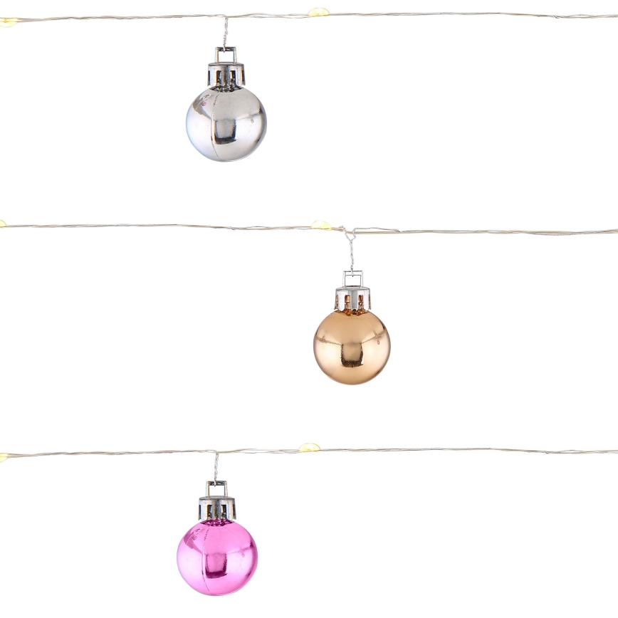 Tri malé gule, strieborná, zlatá a ružová, visia na svetelnom reťazi s malými LED diódami. Gule sú lesklé. Reťaz je na bielom pozadí.