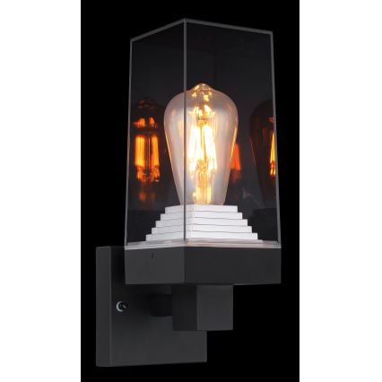 Nástená lampa svieti. Svietidlo má štvorcový tvar, sklenený kryt a vnútri je umiestnená žiarovka Edison. Je pripevnená k stene.