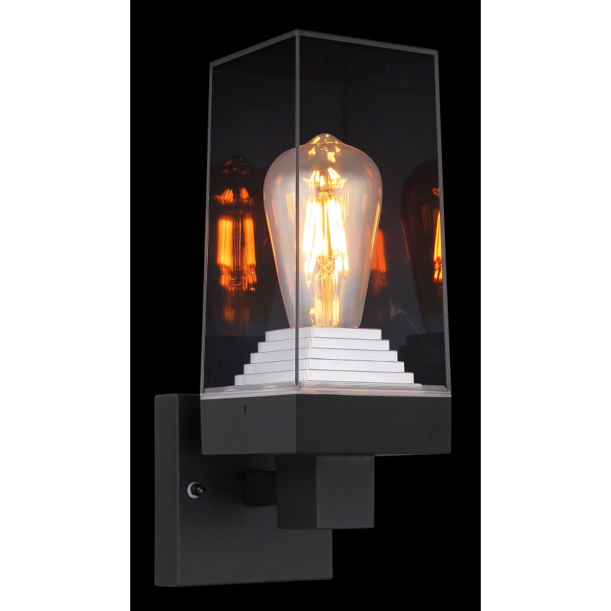 Nástená lampa svieti. Svietidlo má štvorcový tvar, sklenený kryt a vnútri je umiestnená žiarovka Edison. Je pripevnená k stene.