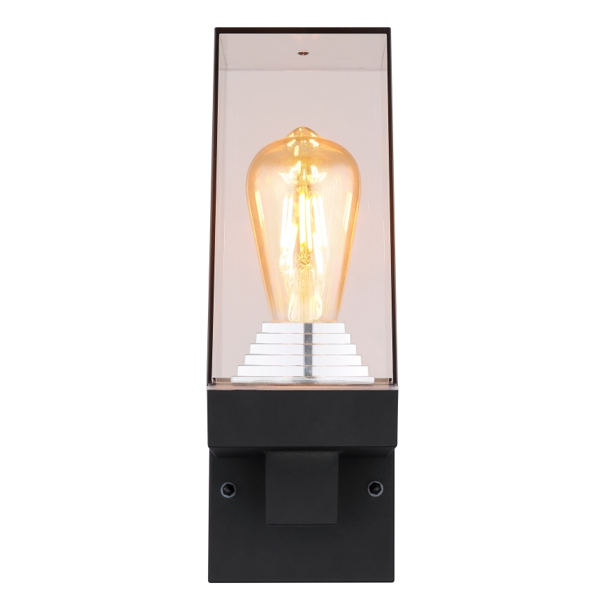 Nástenná lampa svieti. Je umiestnená na stene, v tvare obdĺžnika, s priesvitným sklom a viditeľnou žiarovkou typu Edison.