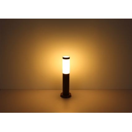 Záhradné osvetlenie svieti. Stojanová lampa s valcovým tienidlom, tmavohnedá, na svetlom povrchu. Svetlo je teplé, žlté.