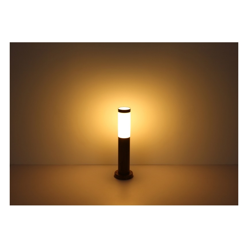 Záhradné osvetlenie svieti. Stojanová lampa s valcovým tienidlom, tmavohnedá, na svetlom povrchu. Svetlo je teplé, žlté.