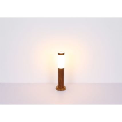 Záhradné svietidlo svieti. Je umiestnené na svetlom podklade. Svietidlo má drevený stĺpik a valcový biely difúzor.
