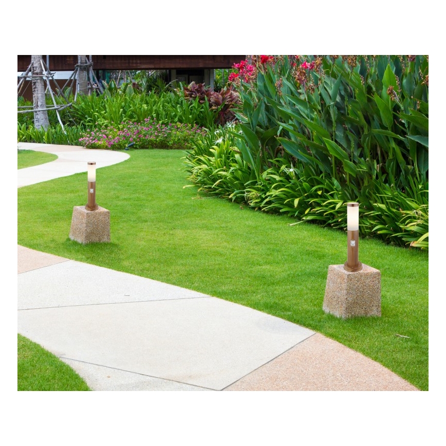 Dve záhradné lampy stoja na betónových podstavcoch v trávniku popri chodníku. Lampy sú umiestnené v zelenej trávnaté ploche lemovanej kvetmi a zeleňou.