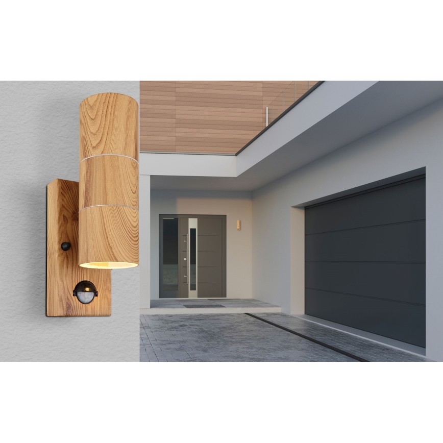 Drevená nástenná lampa s pohybovým senzorom je pripevnená na stene moderného domu. Lampa svieti. V pozadí je viditeľný vstup do domu a garáž.