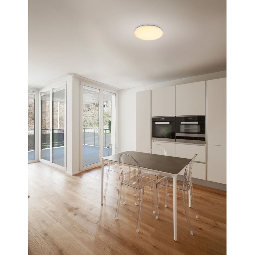 Stropné svietidlo svieti v jedálni. Jedálenský stôl s priesvitnými plastovými stoličkami stojí na svetlej drevenej podlahe. V pozadí je kuchynská linka s dvomi vstavanými rúrami a veľké francúzske dvere vedúce na balkón.