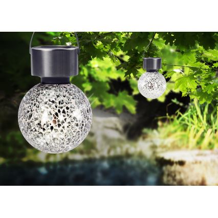 Solárne guľové lampy visia na konároch stromu. Lampy sú z kovu a skla s mozaikovým vzorom. Kontext tvorí zelená záhrada s vodným tokom.