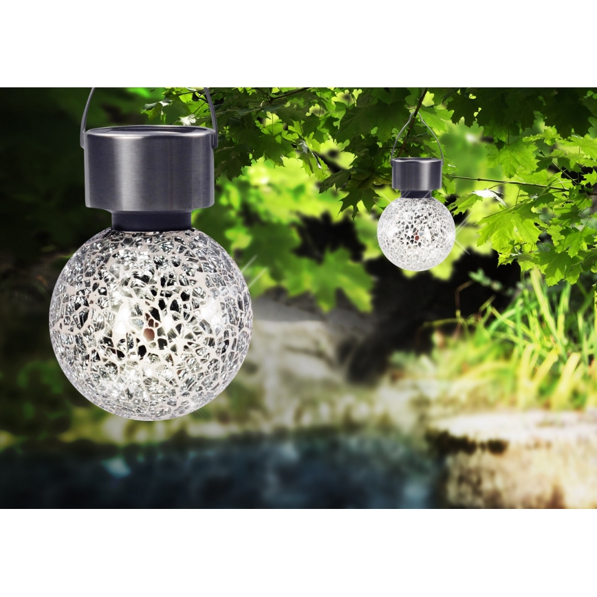 Solárne guľové lampy visia na konároch stromu. Lampy sú z kovu a skla s mozaikovým vzorom. Kontext tvorí zelená záhrada s vodným tokom.