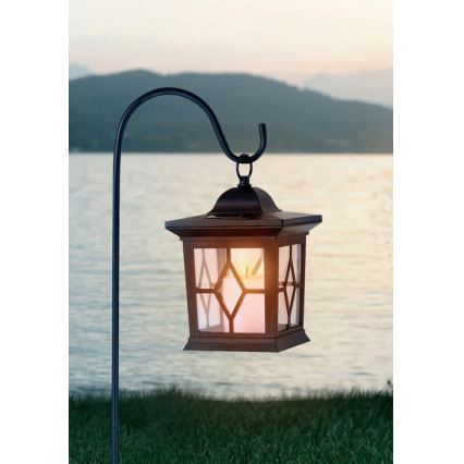 Solárna lucerna visí na záhradnom stojane. Svieti teplým svetlom. Pozadie tvorí vodná plocha a kopce.