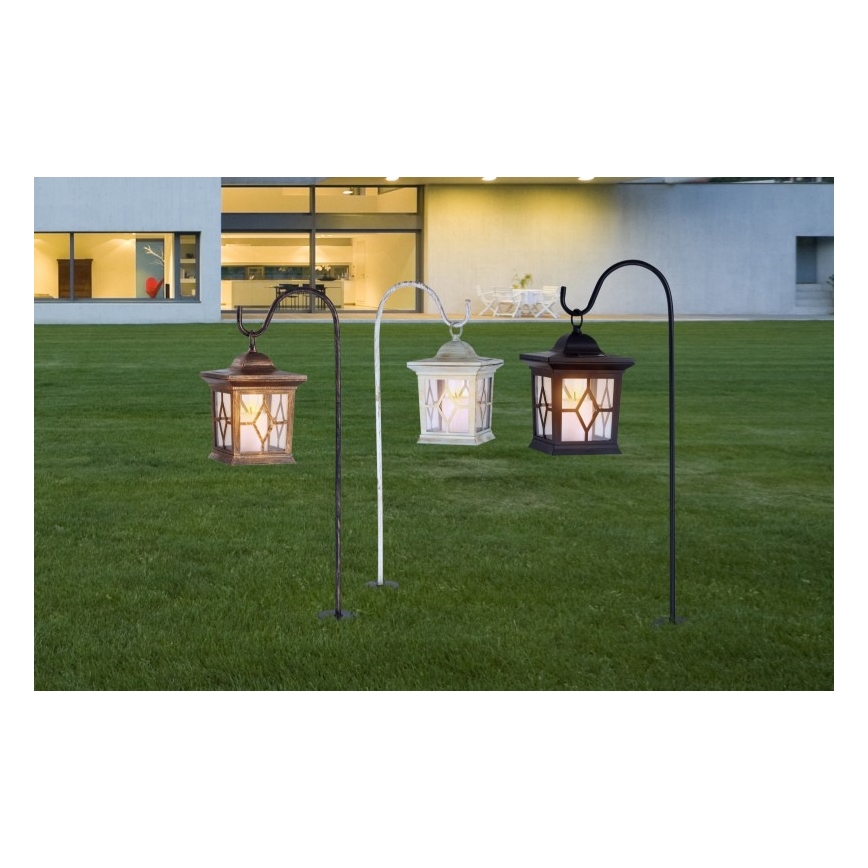 Tri záhradné lampáše visia na zakrivenom stojane. Lampáše sú v hnedo-zlatej, bielej a čiernej farbe. Stojan je umiestnený na zelenom trávniku pred domom.