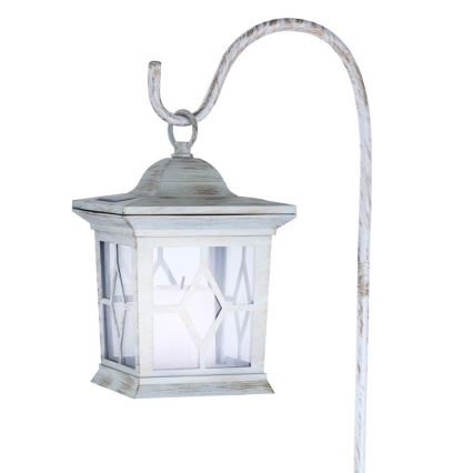 Solárna lampa visí na zahradnom stojane. Lampa je svetlomodrá, štvorcová, s ozdobnými oknami a imitáciou sviečky vo vnútri. Stojan je kovový, zakrivený, svetlomodrej farby.