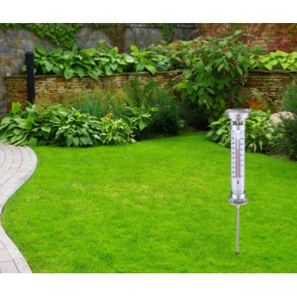 Vonkajší teplomer meria teplotu v trávniku. Nachádza sa v upravenej záhrade s kamenným múrikom a bujnou zeleňou.