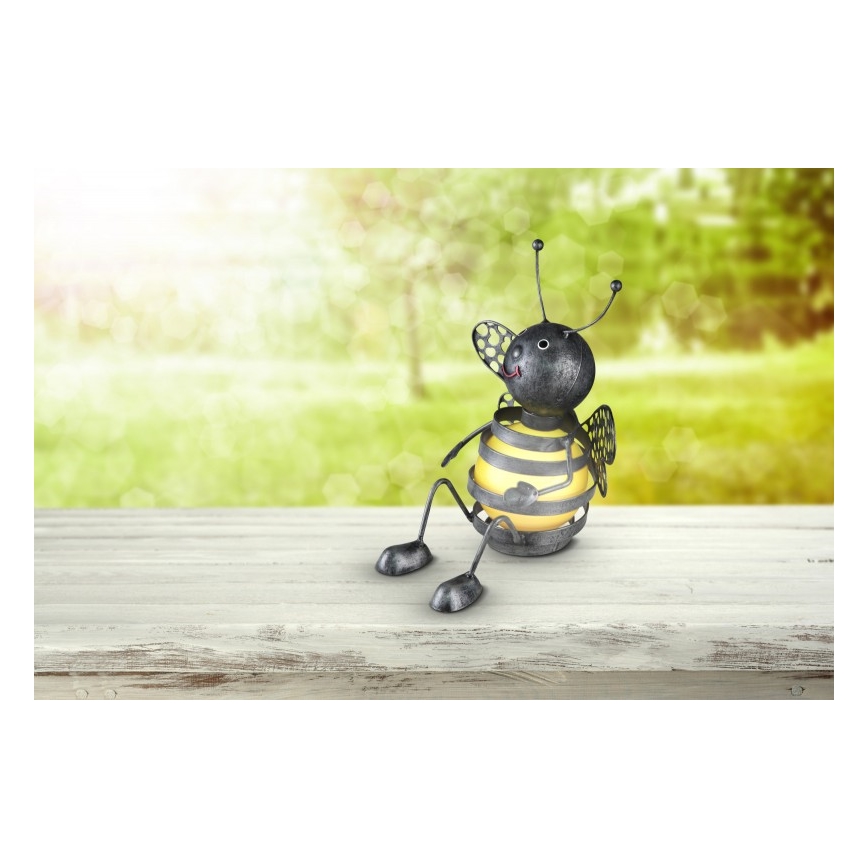 Kovová včielka sedí na drevenom stole. Včielka je tmavosivá, bruško má žlté. Pozadie tvorí rozostrená zelená záhrada.