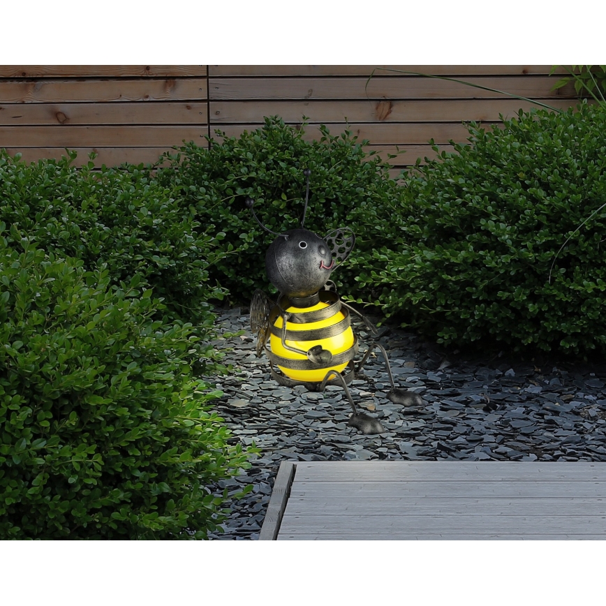 Kovová včela sedí na štrku. Svieti jej bruško. V pozadí je drevený plot a zelené kríky.