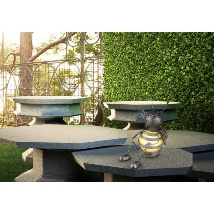 Kovová včela sedí na kamennom stole. Stôl je v záhrade pri nízkom živom plote a kovovej bráne.