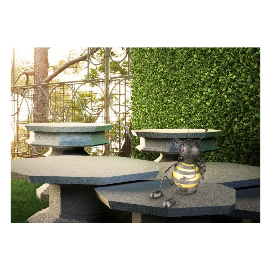 Kovová včela sedí na kamennom stole. Stôl je v záhrade pri nízkom živom plote a kovovej bráne.