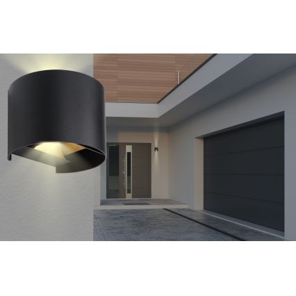 Čierna nástenná lampa svieti. Je umiestnená na stene moderného domu s garážou a vchodom.