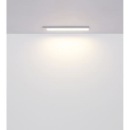 Dlhá úzka LED lampa je pripevnená k stropu a svieti na bielu stenu. Lampa je biela a úzka. Svetlo je jemné a rozsiahle.