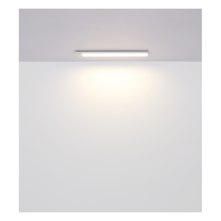 Dlhá úzka LED lampa je pripevnená k stropu a svieti na bielu stenu. Lampa je biela a úzka. Svetlo je jemné a rozsiahle.