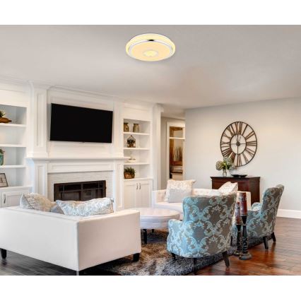 Okrúhle stropné svietidlo s dvojitým kruhovým svetlom svieti v obývacej izbe. Izba obsahuje bielu sedačku, kreslá, krb a televízor.