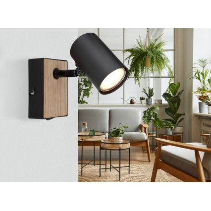 Čierna nástenná lampa s dreveným prvkom svieti. Lampa je umiestnená v obývacej izbe s pohovkou, konferenčnými stolíkmi a izbovými rastlinami.