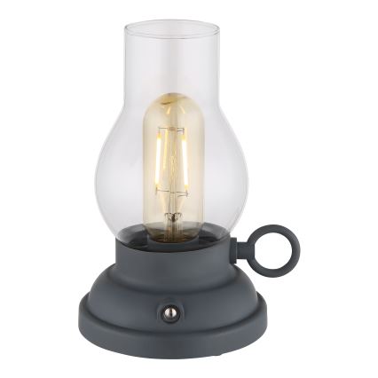 Stolná lampa s tvarom lampáša svieti. Je vyrobená z tmavosivej základne a skleneného obalu, v ktorom je umiestnená LED žiarovka. Lampa má malé madlo.