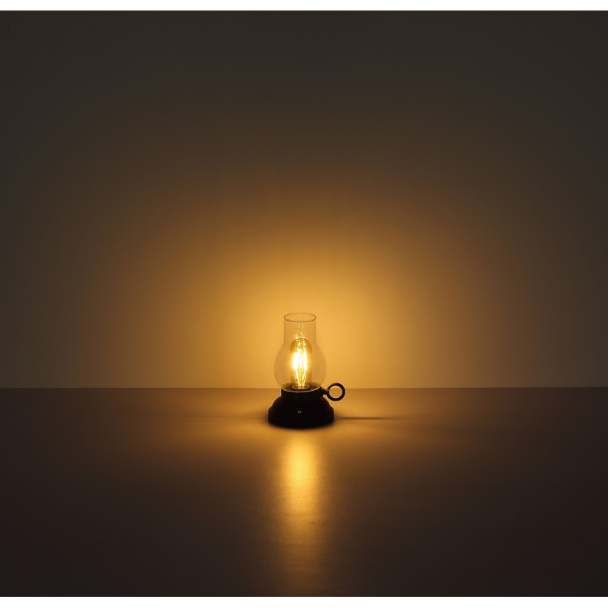 Malá stolová lampa tvarom pripomínajúca petrolejovú lampu svieti na svetlom hnedom povrchu. Lampa je z tmavého kovu a skla, svieti žiarovkou s viditeľným vláknom.