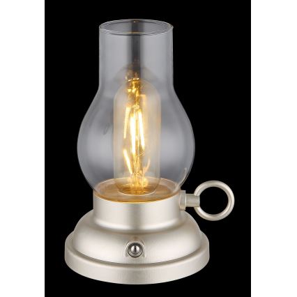 Stolová lampa svieti. Je vyrobená z kovu svetlo béžovej farby a skla. Má tvar petrolejovej lampy s vypínačom na spodnej časti a malým kovový krúžkom na boku.