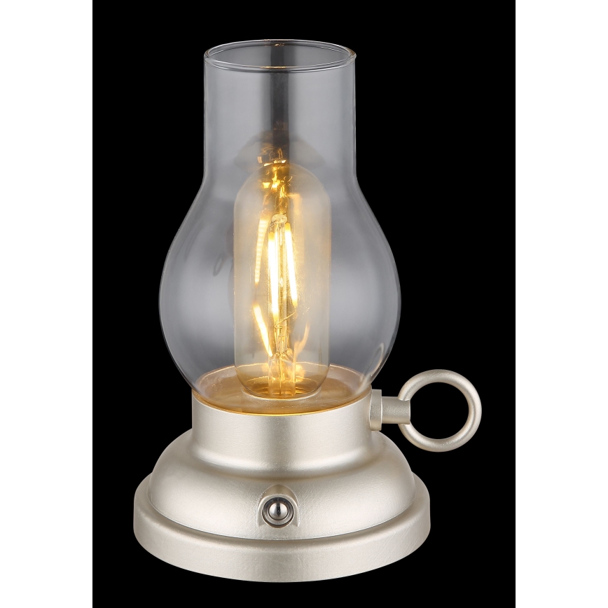 Stolová lampa svieti. Je vyrobená z kovu svetlo béžovej farby a skla. Má tvar petrolejovej lampy s vypínačom na spodnej časti a malým kovový krúžkom na boku.