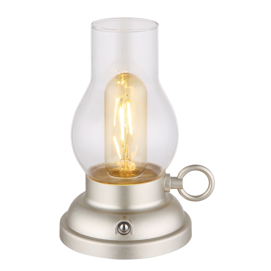 Stolná lampa svieti. Je z kovu svetlo béžovej farby a skla, má tvar petrolejovej lampy. Na spodku má vypínač.