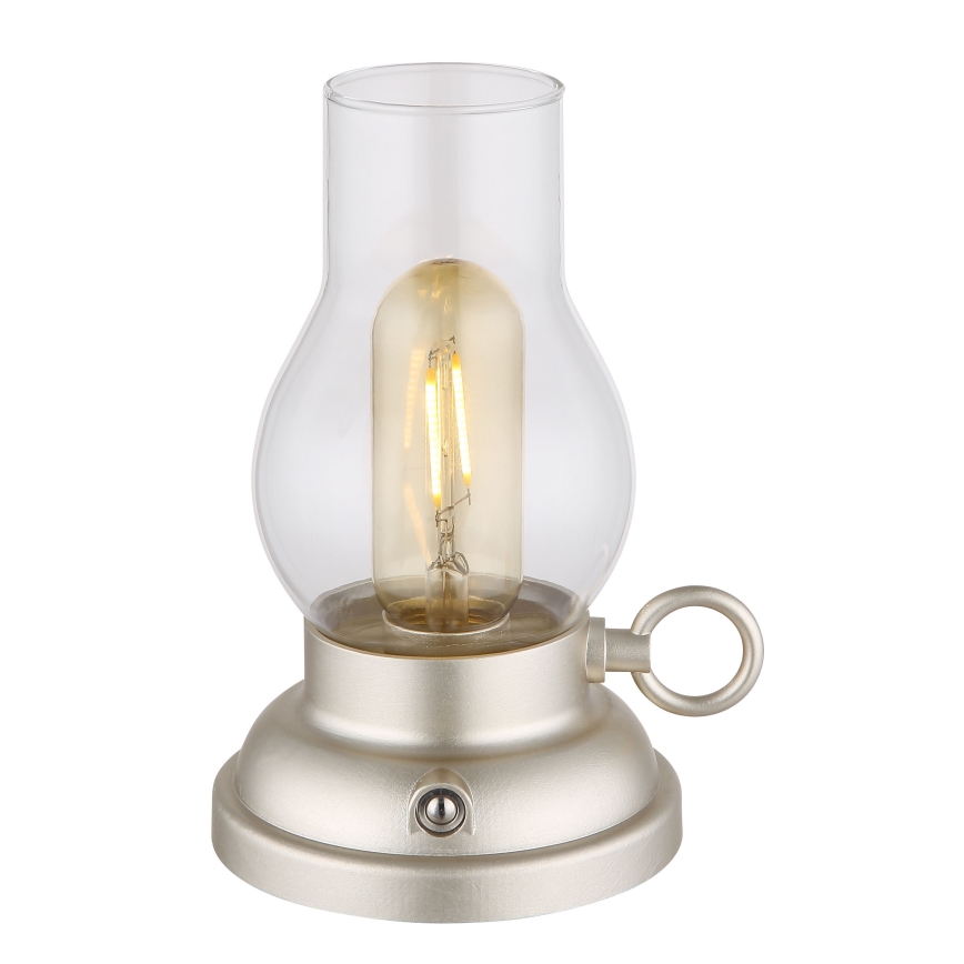 Stolová lampa svieti. Je vyrobená z kovu v svetlo šampanskom odtieni a má sklenený tienidlo v tvare fľaše. Má malý vypínač na spodnej časti.