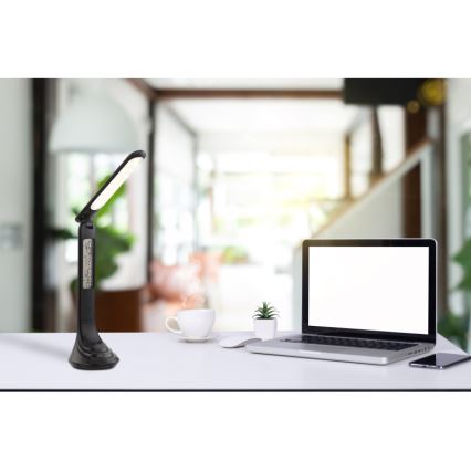 Stolová lampa s LED osvetlením a digitálnym displejom stojí na stole. Vedľa nej je notebook, hrnček a myš. Pozadie tvorí rozostrená miestnosť.