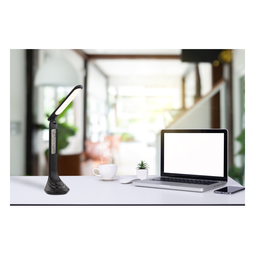 Stolová lampa s LED osvetlením a digitálnym displejom stojí na stole. Vedľa nej je notebook, hrnček a myš. Pozadie tvorí rozostrená miestnosť.