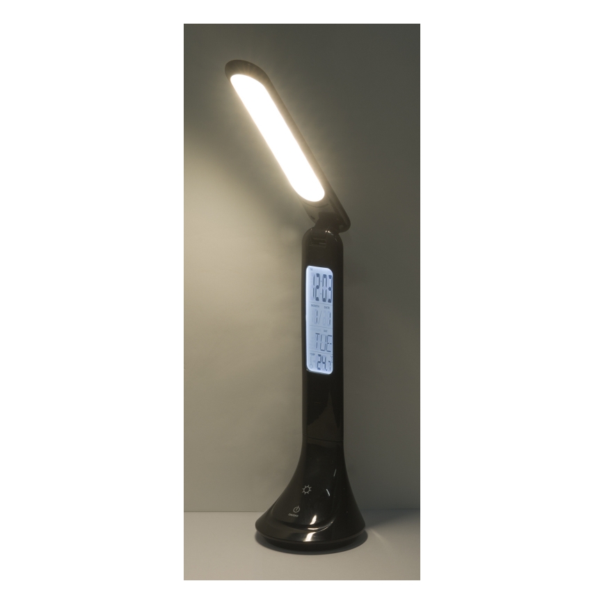 Stolová lampa svieti. Na jej základni je digitálny displej zobrazujúci čas 12:03, utorok a 24h formát. Lampa stojí na stole.