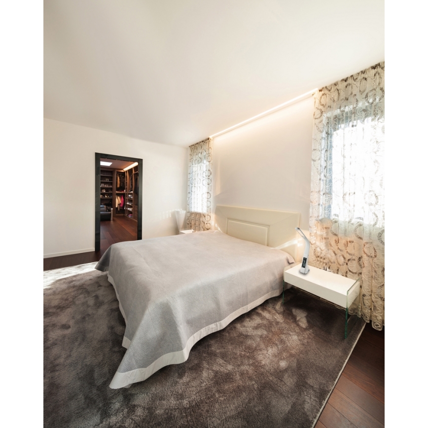 Dvojposteľ leží v spálni na hnedom koberci. Vedľa nej je nočný stolík s nástennou lampou. Za posteľou sú závesy a otvorené dvere vedúce do šatníka.