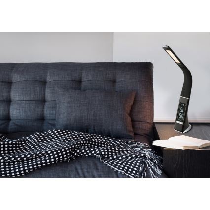 Stolná lampa s budíkom svieti na nočnom stolíku. Lampa je čierna, s digitálnym displejom zobrazujúcim čas a teplotu. Nachádza sa v blízkosti postele so sivou posteľnou bielizňou a čierno-bielou dekou.