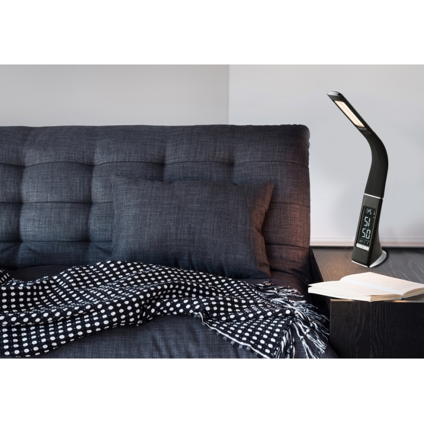 Stolná lampa s budíkom svieti na nočnom stolíku. Lampa je čierna, s digitálnym displejom zobrazujúcim čas a teplotu. Nachádza sa v blízkosti postele so sivou posteľnou bielizňou a čierno-bielou dekou.